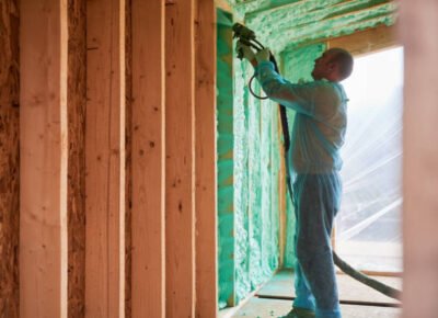 Male builder insulating wooden frame house. Man worker spraying polyurethane foam inside of future cottage, using plural component gun. Construction and insulation concept.